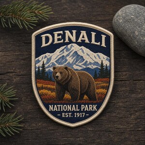 May include: A shield-shaped embroidered patch featuring a brown bear standing in a landscape with a snow-capped mountain, pine trees, and autumn foliage. The text reads DENALI NATIONAL PARK - EST. 1917 - in cream-colored lettering on a dark blue background.
