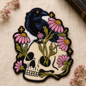 May include: An embroidered patch featuring a black raven perched atop a cream-colored human skull. The design includes pink daisy-like flowers with yellow centers, green stems, and small yellow four-pointed stars, all outlined in thick black thread.