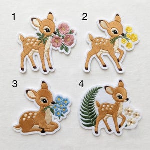 May include: Four embroidered iron-on patches featuring tan fawns with white spots. Patch 1 shows a standing fawn with pink flowers, 2 a standing fawn with yellow flowers, 3 a sitting fawn with blue flowers, and 4 a standing fawn with a green fern.