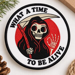 May include: A circular embroidered patch featuring a grim reaper character in a black hooded robe holding a scythe against a red circular background. The text WHAT A TIME TO BE ALIVE is stitched in black along the top and bottom border edges.