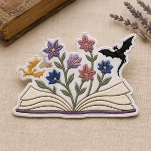 May include: An embroidered patch featuring an open book with pages fanned out. A bouquet of purple, blue, and pink flowers with green stems grows from the center. A small yellow dragon flies on the left, and a black dragon flies on the right.
