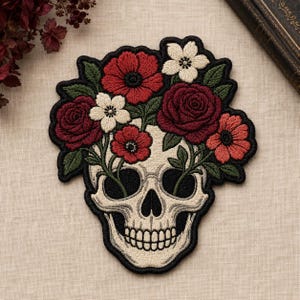 May include: An embroidered patch featuring a cream-colored human skull with a vibrant floral arrangement emerging from the top. The design includes deep red roses, bright red poppies, and small white flowers surrounded by dark green leaves.
