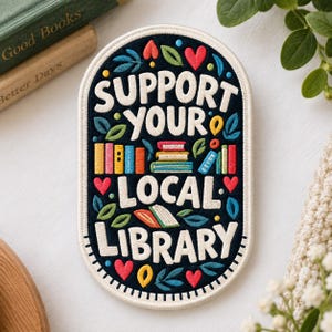 May include: An oval-shaped embroidered patch with a dark navy blue background and a white border. The text reads "SUPPORT YOUR LOCAL LIBRARY" in white letters. The design includes colorful embroidered books, red hearts, and green leaves.
