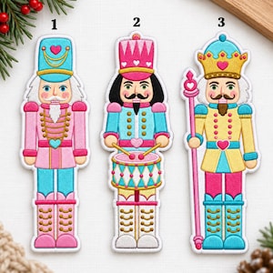 Könnte beinhalten: Drei gestickte Nussknacker-Aufnäher mit den Nummern 1, 2 und 3. Der erste ist rosa und blau, der zweite ist ein Trommler in Rosa und Blau, und der dritte ist ein König in Gelb und Blau mit einem Stab. Alle haben Herzmotive und goldene Verzierungen.