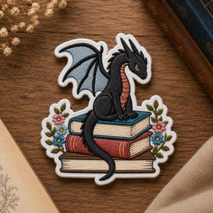 May include: An embroidered patch featuring a black dragon with light blue wings perched atop a stack of three books. The dragon has a tan underbelly. Floral accents in pink and blue flank the books, all outlined in a white border.