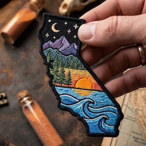 May include: An embroidered patch shaped like the state of California featuring a night sky with a crescent moon and stars above purple mountains, a green pine forest, a golden sunset over the horizon, and blue ocean waves at the bottom.