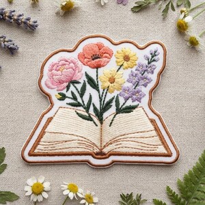 May include: An embroidered patch featuring an open book with pages rendered in cream thread. A bouquet of flowers emerges from the center, including a large pink peony, a coral poppy, two yellow daisies, and a sprig of purple lavender with green stems.