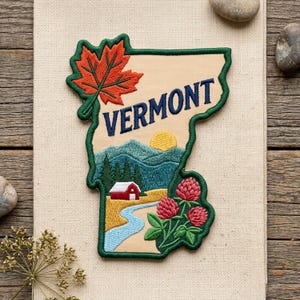 May include: An embroidered patch shaped like the state of Vermont, featuring a red maple leaf, the text VERMONT in dark blue, a yellow sun, green mountains, a red barn, a winding blue river, and pink clover flowers on a tan background.