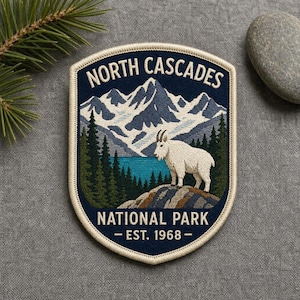 May include: A shield-shaped embroidered patch featuring a mountain goat standing on a rocky outcrop. The design includes a blue lake, green pine trees, and snow-capped mountains under the text NORTH CASCADES NATIONAL PARK - EST. 1968 -.