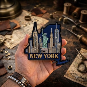 May include: A dark blue embroidered patch shaped like the state of New York, featuring gold and light blue thread depictions of the Statue of Liberty and iconic skyscrapers. The text NEW YORK is stitched in gold at the bottom of the patch.
