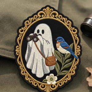 May include: An oval embroidered patch featuring a white ghost holding black binoculars and wearing a brown satchel. A blue and orange bird sits on a green leafy branch next to a white flower. The scene is enclosed in an ornate gold frame.