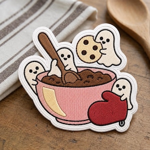 May include: An embroidered patch featuring three small white ghosts interacting with a pink bowl of brown batter. One ghost holds a brown wooden spoon, another holds a chocolate chip cookie, and the third ghost rests against a red oven mitt.