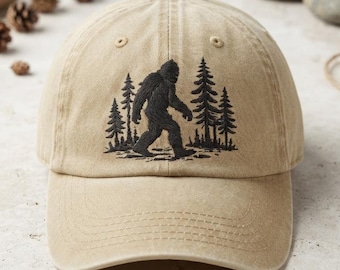 Può includere: Un cappellino da baseball color marrone chiaro con un ricamo nero che raffigura la sagoma di un Bigfoot che cammina attraverso una foresta di pini. Il cappellino presenta una corona strutturata, una visiera curva e occhielli cuciti.