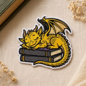 May include: An embroidered patch featuring a yellow dragon with black-outlined wings sleeping on top of two stacked dark gray books. The dragon has small horns and a curved tail, with white thread bordering the entire shape of the design.