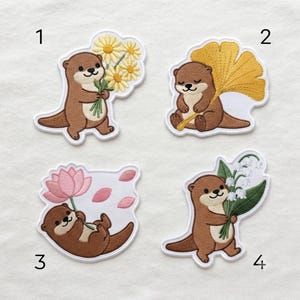 May include: Four embroidered patches featuring brown otters with cream-colored bellies. Patch 1 shows an otter holding yellow daisies. Patch 2 shows an otter with a yellow ginkgo leaf. Patch 3 shows an otter with a pink lotus. Patch 4 shows an otter with lily of the valley.