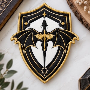 May include: A shield-shaped embroidered patch featuring a black dragon with gold wings centered on a white background. The design is framed by a thick gold border and includes four small gold stars in the upper corners and a star at the base.