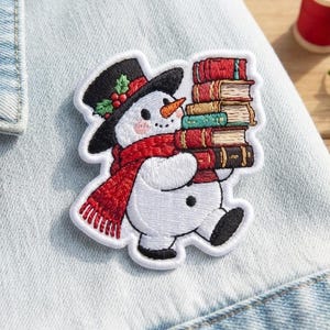 May include: An embroidered patch featuring a white snowman wearing a black top hat with holly and a red scarf, holding a stack of five colorful books. The snowman has a carrot nose, rosy cheeks, and black buttons on a light blue denim fabric.