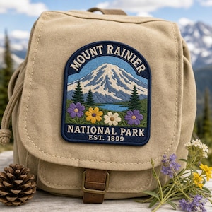May include: A tan canvas backpack featuring an embroidered patch of Mount Rainier National Park. The patch displays a snow-capped mountain, pine trees, and colorful wildflowers with the text MOUNT RAINIER NATIONAL PARK EST. 1899.