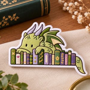 May include: An embroidered patch featuring a light green dragon with lavender horns resting its head and front paws on a row of colorful, upright books. The books are decorated with various patterns including flowers, hearts, and geometric lines.