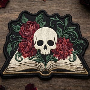 May include: An embroidered patch featuring an open book with a cream-colored skull resting on the pages. The design includes deep red roses and a dark red dahlia, surrounded by green swirling vines and leaves, all set against a black background.