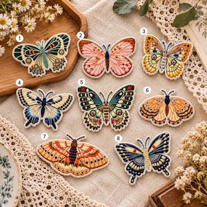 May include: Eight embroidered butterfly patches labelled 1 through 8, featuring diverse colour palettes including green, pink, yellow, blue, and orange. Each patch displays intricate thread work and distinct wing patterns on a light fabric background.