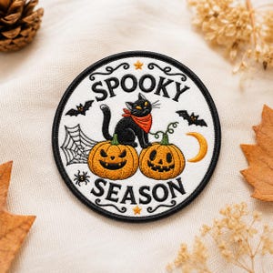May include: A circular embroidered patch with a white background and black border. It features a black cat wearing an orange bandana, two orange jack-o'-lanterns, a spider web, a spider, two bats, a yellow crescent moon, and two stars. The text reads SPOOKY SEASON.
