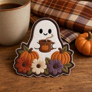May include: An embroidered patch featuring a white ghost holding a brown mug with a pumpkin design. The ghost is surrounded by two orange pumpkins, a rust-colored flower, a white flower, and a purple flower, all outlined in dark brown thread.