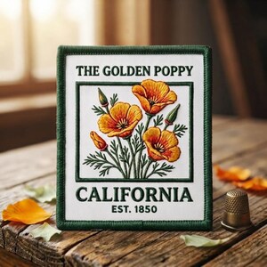 May include: A rectangular embroidered patch with a dark green border on a white background. It features orange poppy flowers and green stems. The text reads THE GOLDEN POPPY CALIFORNIA EST. 1850 in dark green lettering. A metal thimble sits nearby.