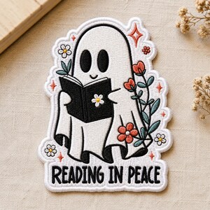 May include: An embroidered patch featuring a white ghost with black eyes reading a black book. The design includes orange and white flowers, green leaves, and small orange star accents. The text "READING IN PEACE" is stitched in black at the bottom.