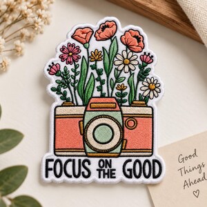 May include: An embroidered patch featuring a coral-colored camera with a mint green lens, topped with a bouquet of colorful flowers including pink, orange, and white blossoms. The text "FOCUS ON THE GOOD" is stitched in black at the bottom.