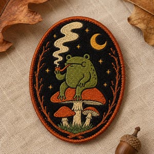 May include: An oval embroidered patch featuring a green frog sitting on a large orange mushroom, smoking a pipe with swirling smoke. The background is black with a yellow crescent moon and stars, framed by a rust-colored border with branch details.