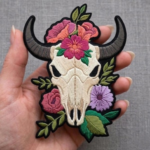 May include: An embroidered patch featuring a cream-colored animal skull with dark gray horns, surrounded by vibrant pink, orange, and purple flowers with green leaves, all outlined in a thick black border against a gray background.