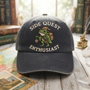 Könnte beinhalten: Eine anthrazitfarbene Baseballkappe mit einer Stickerei eines grünen Frosches, der einen Hut trägt, einen Korb auf dem Rücken hat und eine kleine Uhr hält. Der Text "SIDE QUEST ENTHUSIAST" ist in Weiß über und unter dem Frosch gestickt.