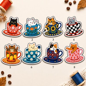 May include: Eight embroidered iron-on patches featuring cats sitting in various patterned teacups. Designs include a black cat in a red floral cup, a white cat in a blue floral cup, a tabby in a sunflower cup, and a gray cat in a checkered cup.