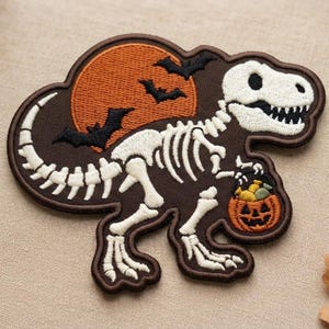 May include: An embroidered patch featuring a white T-Rex skeleton on a dark brown background. The design includes a large orange full moon with black silhouettes of flying bats and the dinosaur holding a small orange jack-o'-lantern bucket.