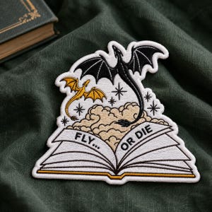 May include: An embroidered patch featuring an open book with the text FLY... OR DIE. A large black dragon with wings spread emerges from the pages above beige clouds, while a smaller gold dragon flies to the left. Black star icons surround them.