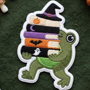 Può includere: Una toppa ricamata raffigurante una rana verde che trasporta una pila di libri sormontata da un cappello da strega nero. I libri sono decorati con una zucca, una luna crescente, un pipistrello e un fantasma. La toppa ha un bordo bianco.