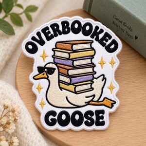 May include: An embroidered patch featuring a white goose wearing black sunglasses, carrying a tall stack of colorful books on its back. The text OVERBOOKED GOOSE is stitched in black above and below the goose, surrounded by small gold stars.