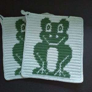 Könnte beinhalten: Zwei quadratische, gehäkelte Topflappen. Jeder Topflappen zeigt ein grünes Froschdesign auf hellgrünem Hintergrund. Die Topflappen haben eine Schlaufe zum Aufhängen. Handgemachte Küchendekoration.