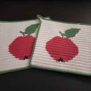 Könnte beinhalten: Zwei weiße gehäkelte Topflappen mit grünen Blättern und roten Äpfeln. Die Topflappen haben einen grünen Rand.