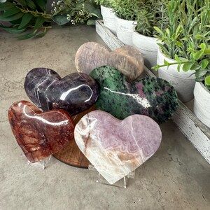 May include: A selection of heart-shaped stones in shades of purple, green, and red. The stones are presented on clear stands, highlighting their individual patterns and textures. Decorative items.