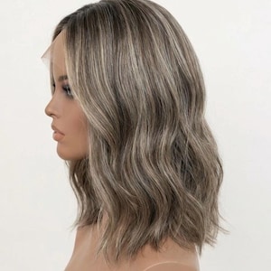 Op de afbeelding: Een bob-stijl pruik met een kanten voorkant, met een mix van asbruine en blonde highlights. Het haar heeft een golvende textuur en is gestyled om het gezicht te omlijsten. De pruik is ontworpen om er natuurlijk uit te zien.