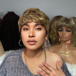 Op de afbeelding: Een persoon draagt een grijze trui met een gouden kettingketting en meerdere ringen aan de vingers. De persoon draagt een kort, blond pruik met een scheiding aan de zijkant. De persoon staat voor twee mannequins met pruiken.