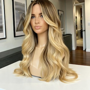 Könnte beinhalten: Blonde Perücke mit langem, gewelltem Haar, präsentiert auf einem Schaufensterkopf. Die Perücke hat dunkle Wurzeln mit helleren blonden Highlights und einen natürlich aussehenden Haaransatz. Das Haar fällt in weichen Wellen und erzeugt einen voluminösen und stilvollen Look.