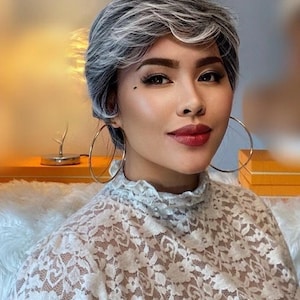 Op de afbeelding: Een vrouw met kort, zilvergrijs haar die grote creolen draagt en een wit kanten topje.