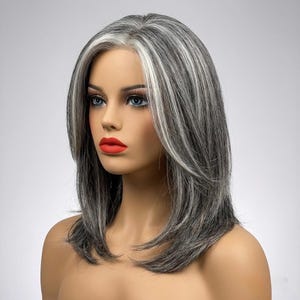 Op de afbeelding: Schouderlange pruik met een gelaagde snit. De pruik heeft grijze en witte lokken, met een prominente witte streep aan de voorkant. De mannequin draagt rode lippenstift en heeft blauwe ogen.