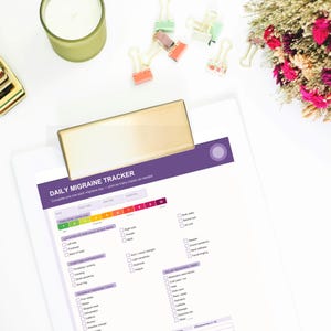May include: A white clipboard with a gold clip holds a "Daily Migraine Tracker" form. A gold stapler, a green candle, and colorful binder clips are scattered around. Dried flowers with pink and orange hues are also present.