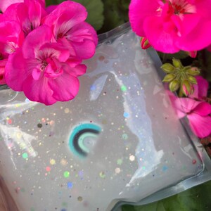 Könnte beinhalten: Ein durchsichtiger Plastikbeutel gefüllt mit weißem Schleim mit schimmerndem Glitzer und einem Regenbogenmuster. Der Beutel ist von rosa Blumen umgeben.