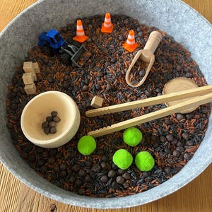 Könnte beinhalten: Eine sensorische Schale gefüllt mit schwarzem und braunem Reis, kleinen Holzblöcken, einem blauen Spielzeug-LKW, orangefarbenen Verkehrskegeln, einem Holzschöpfer und grünen Pompons. Eine Holzschale enthält kleine braune Bohnen, und Holzpinzetten sind ebenfalls enthalten.