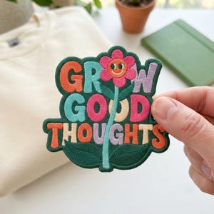 May include: An embroidered patch featuring the text GROW GOOD THOUGHTS in multi-coloured block letters. A pink flower with a smiling face serves as the letter O in GROW. The patch has a dark green background with a stem and leaves at the base.
